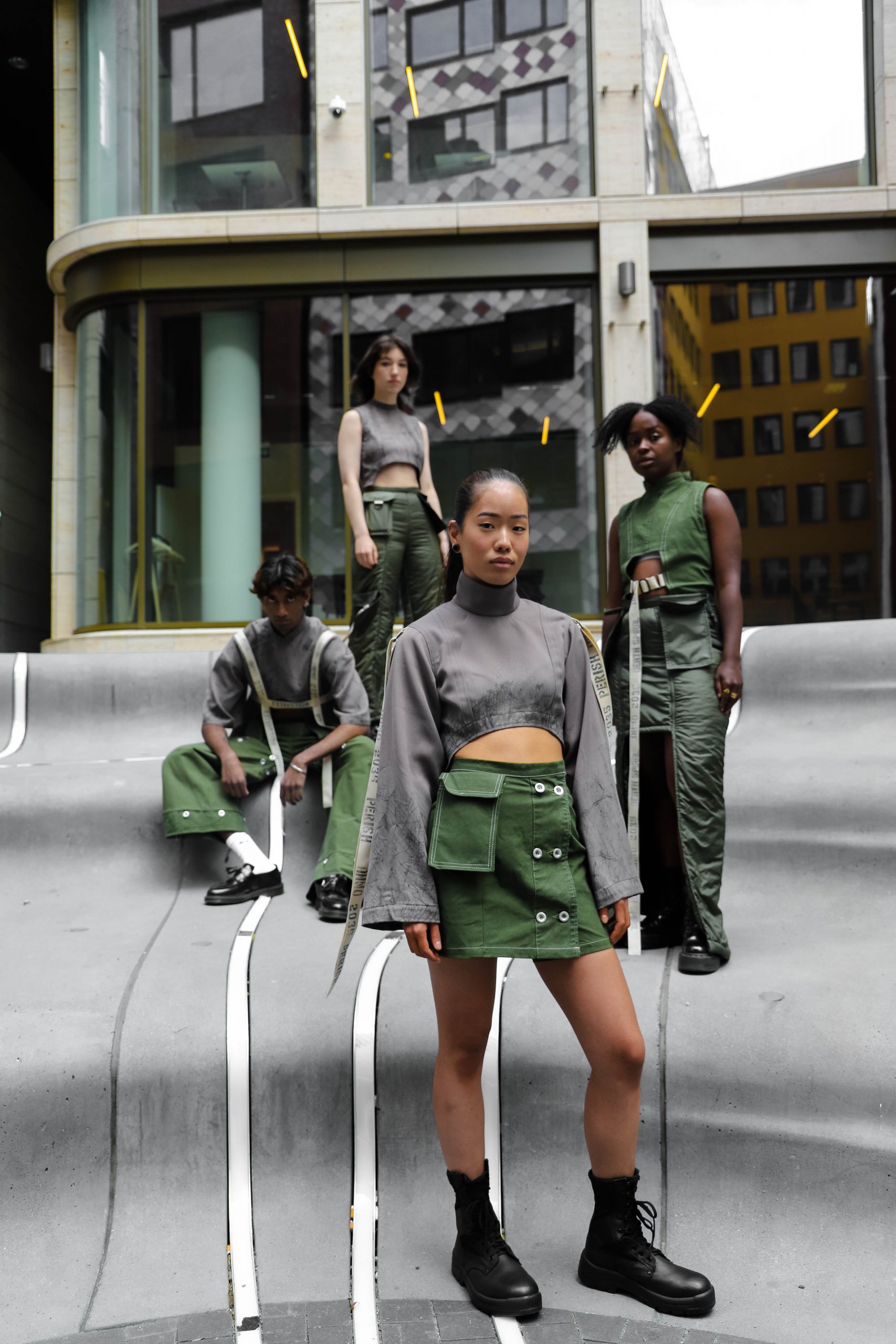 Group photo, four people standing different distance to viewer wearing cropped tops in green and grey with white straps/belts and green skirts and pants with pocket and button details.
