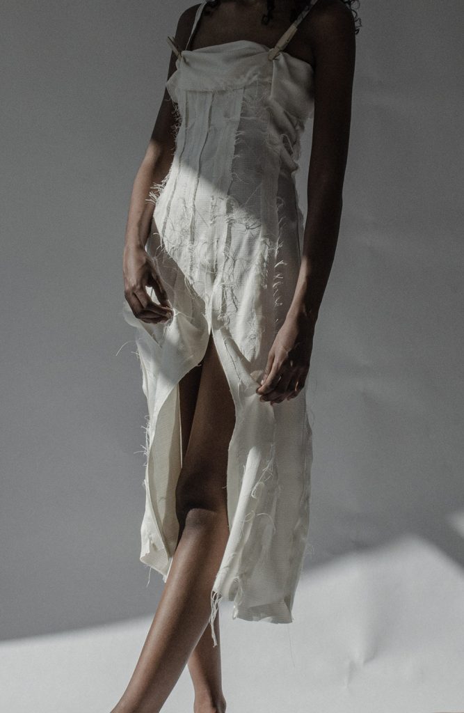 En kvinne er fotografert fra skuldrene og ned mot en lys bakgrunn. Hun har på seg en hvit kjole med sømdetaljer som har splitt langs det ene beinet.