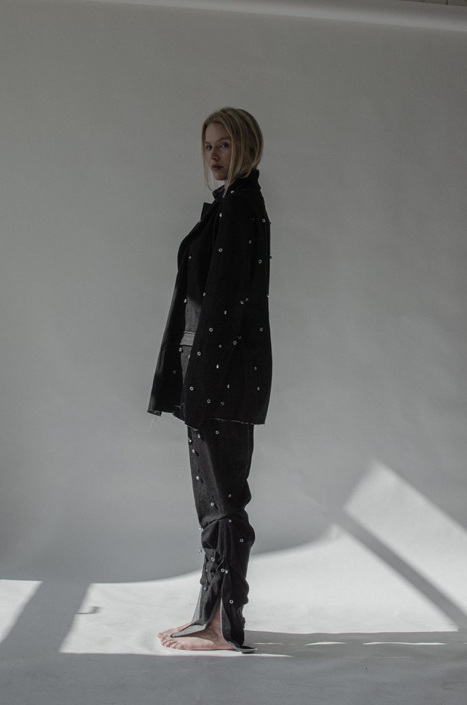 En kvinne er fotografert barbeint i helprofil mot en lys bakgrunn. Hun ser i kamera og er kledd i et mørkt sett bestående av en jakke og bukse-kombinasjon med perledetaljer.