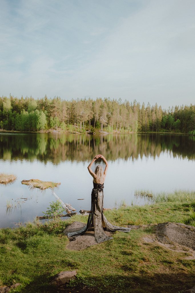 En eldre kvinne står foran et vann i skogen. Hun holder hendene over hodet og ser utover vannet. Kjolen hennes er lang og ligger utover gresset og berget.