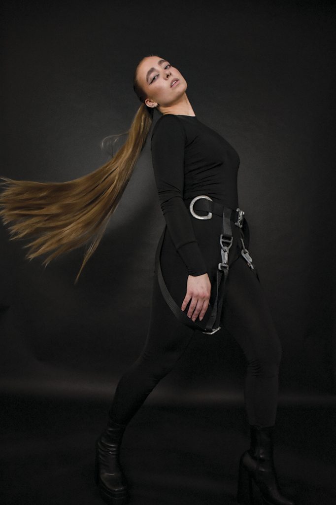En kvinne står fotografert mot en mørk bakgrunn i sorte klær og combat boots. Hun har langt hår som er fotografert i bevegelse bak henne, mens hun ser i kamera. Undertøyet har ulike detaljer inspirert av motorsykkel- og pilotklær, som sikringsseler