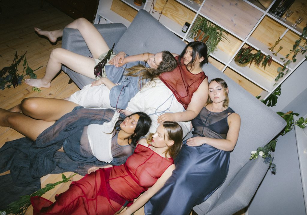 Fem kvinner er fotografert i fugleperspektiv mens de ligger på en sofa som står foran en bokhylle. Kvinnene lener seg mot hverandre og ligger henslengt med lukkede øyne. Det ligger blomster rundt dem, og på seg har de klær i materialer i hvite, blå- og rødtoner.