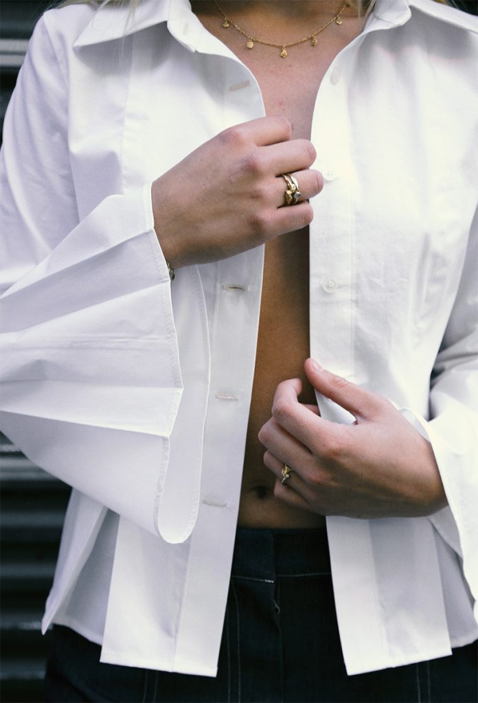 Et detaljbilde av en hvit skjorte med foldedetaljer på ermene. Skjorten er ukneppet og stylet mot en mørk bukse og gull-smykker.