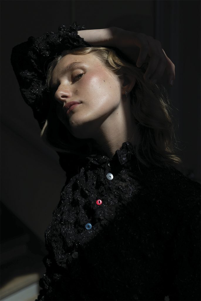 En kvinne er fotografert i chiaroscuro-stil mot mørk bakgrunn. Lyset treffer deler av ansiktet hennes og fremhever materialet i den sorte toppen og knappene i ulike farger.