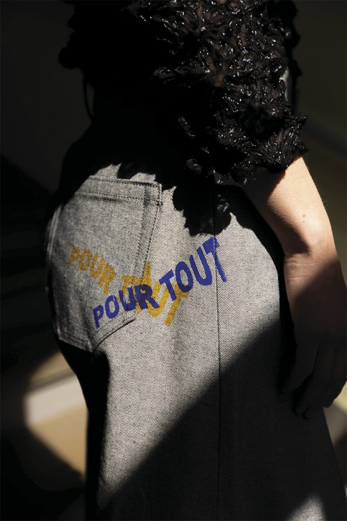 Detaljbilder av baksiden av grå bukser med teksten "POUR TOUT" trykket i blått og gult. Det er sterk kontrast hvor lyset treffer buksene og fremhever detaljen.